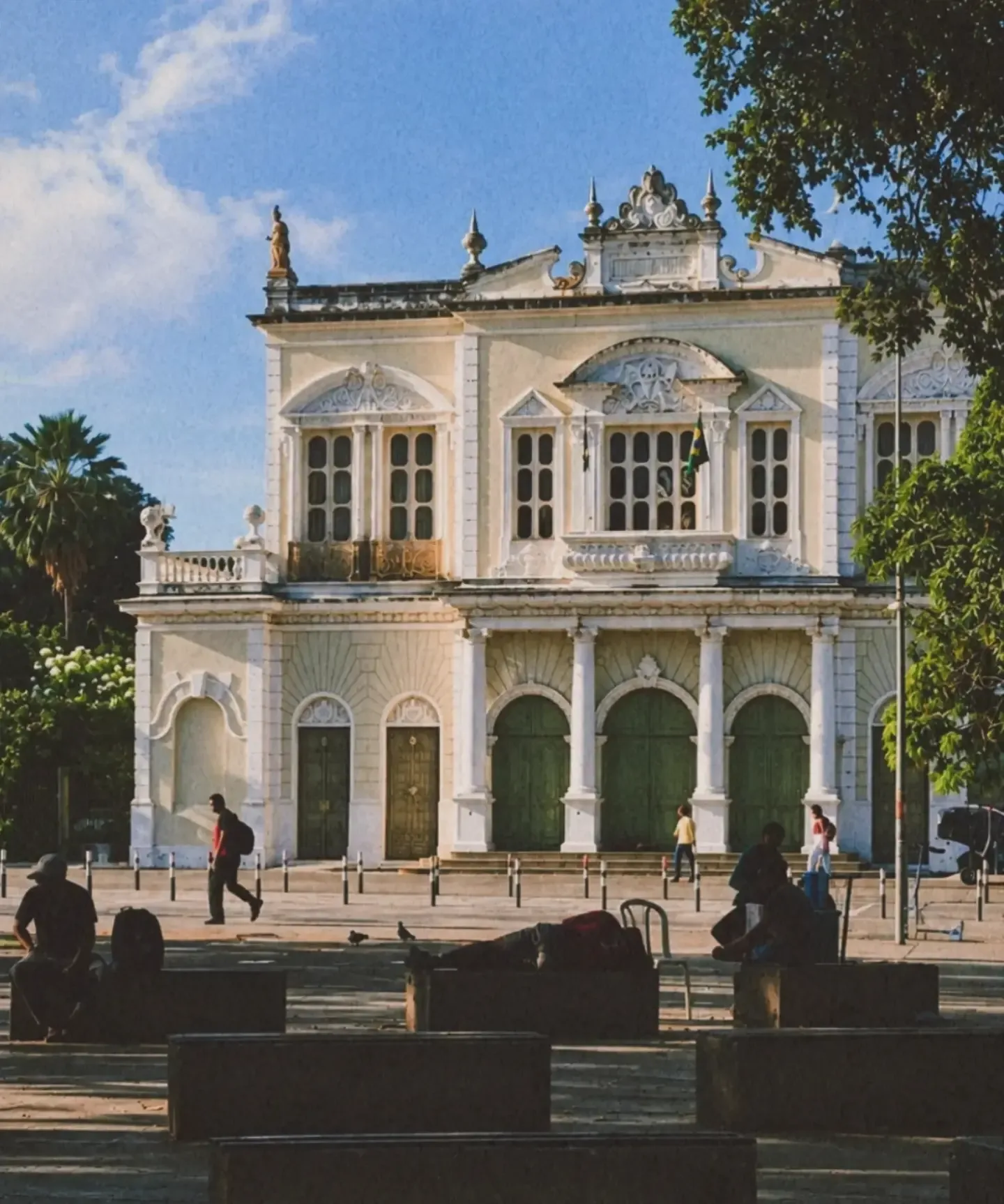 Edifício branco de estilo clássico com portas verdes e uma praça em frente, sob céu azul.