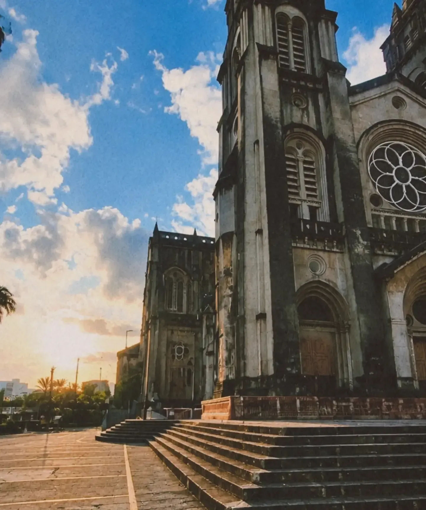 Lateral de uma igreja de pedra com torres góticas e escadaria, ao pôr do sol, sob céu azul com nuvens.