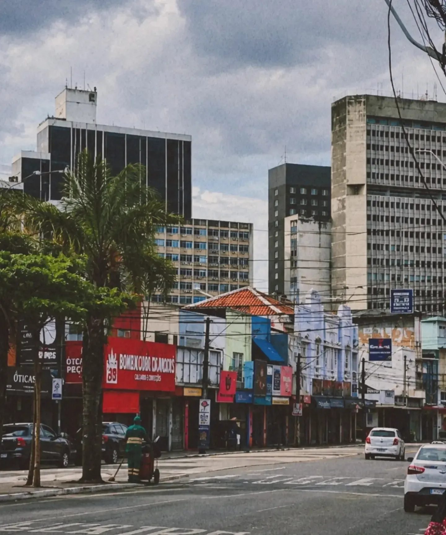 Rua do centro com prédios, fachadas coloridas, carros e uma palmeira, sob céu parcialmente nublado.
