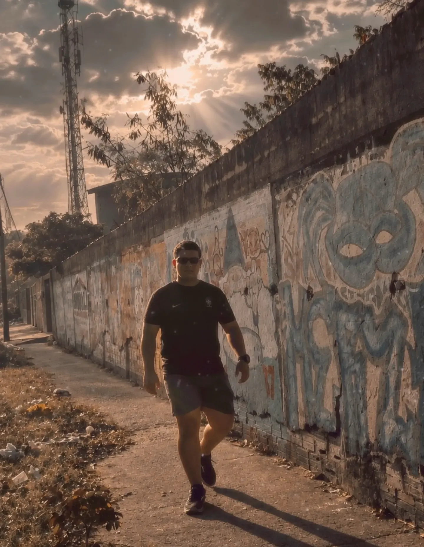 Homem de camiseta preta e bermuda caminhando em uma calçada ao lado de um muro grafitado, com o sol atrás das nuvens.