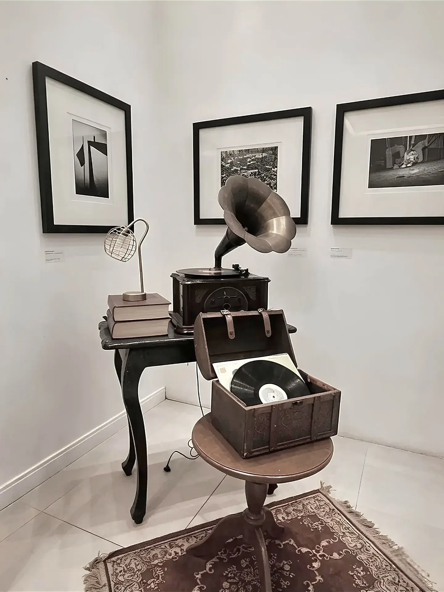 Canto de uma galeria com quadros em preto e branco na parede, um gramofone com disco e uma mesa de madeira sobre um tapete.